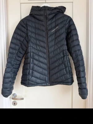 Svart dunjacka med huva från Peak Performance - Snygg svart dunjacka från Peak Performance med huva och dragkedja framtill. Jackan har ett quiltat mönster och är lätt vadderad för extra värme. Perfekt för kalla vinterdagar och har en stilren, sportig look.