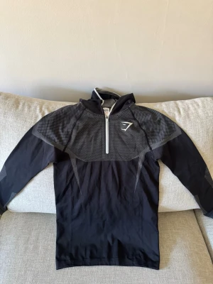 Gymshark onyx V5 hoodie - Snygg svart långärmad träningströja från Gymshark med halv dragkedja framtill och gråmelerade detaljer över axlar och armar. Tillverkad i ett stretchigt funktionsmaterial som andas, perfekt för gymmet eller löprundan. Inte använd bara tagit ut ur originalförpackningen.