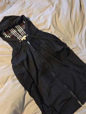 Svart hoodie från Burberry - Svart hoodie från Burberry med dragkedja och klassiskt rutigt foder i huvan. Märkets logga är diskret broderad på bröstet. Hoodien har fickor framtill och justerbar dragsko i huvan. Perfekt för en stilren och avslappnad look.