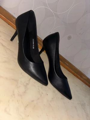 Svarta klassiska pumps med smal klack - Säljer ett par svarta pumps med spetsig tå och smal klack. Skorna har en stilren och tidlös design i slätt material som ger en elegant look. Perfekta för att lyfta vilken outfit som helst. De är oanvända original priset är 449 säljer de för 249kr
