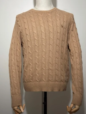 Beige kabelstickad tröja Polo Ralph Lauren - Kabelstickad beige tröja från Polo Ralph Lauren med rund halsringning och diskret broderad logotyp på bröstet. Tröjan har ribbade muddar vid ärmslut och nederkant och är tillverkad i mjukt material, perfekt för lager på lager.