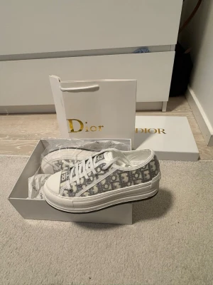 Dior B23 grå sneakers - Snygga Dior B23 sneakers i lågt snitt med grått och vitt monogrammönster. Skorna har vita snören, gummisula och detaljer med 'Christian Dior' på plösen. Ovandelen är i canvas och mesh med klassisk Dior-logga över hela skon. Perfekta för dig som gillar exklusiva streetwear-vibbar.   