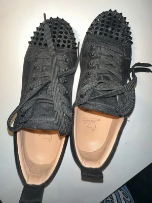 Svarta spikes-sneakers Christian Louboutin - Unika svarta sneakers från Christian Louboutin med mattsvarta spikes på tån och klassisk röd sula. Skorna har snörning, ovandel i mocka och en låg silhuett. Perfekta för dig som vill sticka ut med en edgy och exklusiv stil.