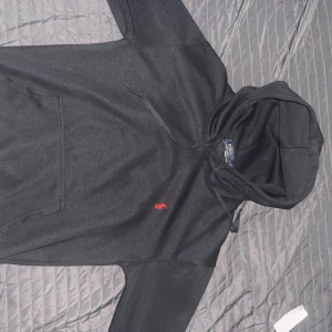 Svart hoodie från Polo Ralph Lauren - Svart hoodie från Polo Ralph Lauren med klassisk röd broderad logga på bröstet. Hoodien har dragsko i huvan och en stor magficka. Perfekt för en chill och stilren look.
