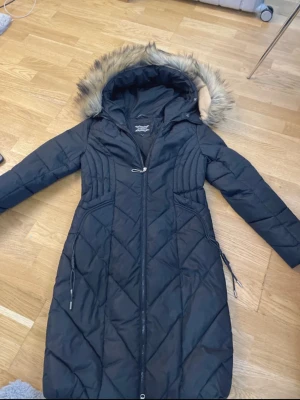 Svart lång dunjacka med fuskpäls - Snygg svart lång dunjacka från Everest med quiltad design och huva med fuskpäls. Jackan har dragkedja framtill, långa ärmar och praktiska fickor med dragkedja. Perfekt för kalla vinterdagar och riktigt varm.