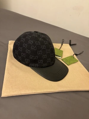 Svart keps från Gucci med GG-mönster - Snygg svart keps från Gucci med klassiskt GG-monogram över hela tyget och böjd skärm i svart läder. Kepsen har en exklusiv känsla och är perfekt för dig som vill sticka ut med en lyxig accessoar.