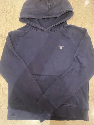 Mörkblå hoodie från GANT - Mörkblå hoodie från GANT med klassisk huva och liten broderad logga på bröstet. Tillverkad i mjukt bomullsmaterial och har en känguruficka framtill. Perfekt för en avslappnad och stilren look.