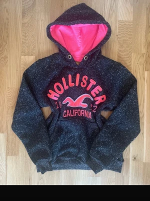 Mörkgrå Hollister hoodie med rosa huva - Snygg mörkgrå hoodie från Hollister med rosa insida på huvan och stora rosa tryck på bröstet. Klassisk känguruficka framtill och ribbade muddar. Perfekt för dig som gillar streetwear och vill ha något bekvämt och stilrent.