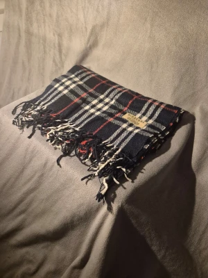 Rutig halsduk från Burberry - Mörkblå halsduk från Burberry med klassiskt rutmönster i vitt och rött. Halsduken har fransar i ändarna och en tygpatch med Burberry-loggan. Materialet ser ut att vara ull, vilket gör den både mjuk och varm. Perfekt för dig som gillar tidlös design.