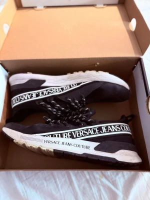 Svarta Versace Jeans Couture sneakers - Snygga svarta sneakers från Versace Jeans Couture med vita detaljer och logga längs sidan. Skorna har svartvitrandiga skosnören och en tjock vit sula. Materialet är en mix av textil och syntet. Perfekta för dig som vill ha en trendig och sportig look.