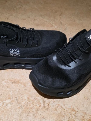 Svarta On x Loewe sneakers - Snygga svarta sneakers från On x Loewe med stickad ovandel och unika detaljer. Skorna har elastiska svarta snören, diskret logga på sidan och en chunky sula med hål för extra stil. Perfekta för dig som gillar modern och sportig design.