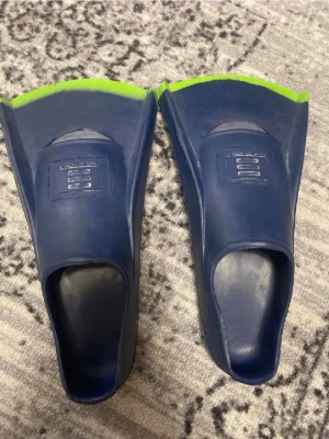 Blå simfenor med gröna detaljer - Säljer ett par blå simfenor med gröna kanter, perfekta för simträning eller snorkling. Tillverkade i gummi för bra komfort och grepp. Märkta med storlek 40-41 och tillverkade i Malaysia. Snygg och sportig design som passar dig som vill ta simningen till nästa nivå.