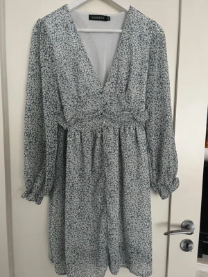 Blommig v-ringad klänning Fashion - Söt klänning med småblommigt mönster i vitt och grått från Fashion. Klänningen har v-ringning, långa puffärmar med resår vid ärmslut och markerad midja. Lätt och luftig modell som slutar ovanför knäna. Perfekt för en trendig look. Storlek L, men passar mig som har S.