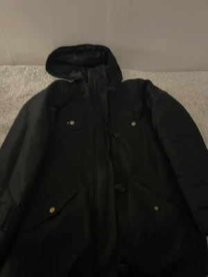 Svart parkasjacka med huva - Snygg svart parkasjacka med huva och guldfärgade knappar. Jackan har flera fickor framtill och justerbara ärmslut. Perfekt för kalla dagar och har en klassisk, stilren look som passar till det mesta.