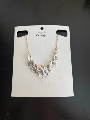 Gnistrande halsband från George - Snyggt halsband från George med gnistrande, klara stenar i olika former som bildar ett blommigt mönster framtill. Kedjan är tunn och guldfärgad, vilket ger en elegant touch. Perfekt för dig som vill sticka ut med lite extra bling.