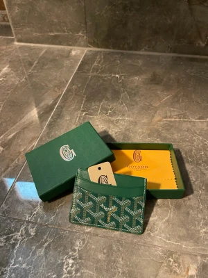 Grön Plånbok - Snyggt grönt plonbok med klassiskt mönster i vitt och mörkgrönt. Tillverkat i exklusivt läder och canvas, perfekt för kort och ID. Kommer med original ask och putsduk. Stilren och lyxig accessoar för dig som gillar designer.