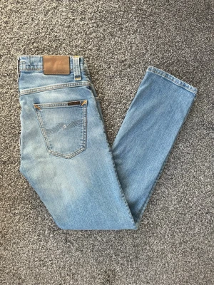 Ljusblå slim jeans från Nudie - Säljer ett par ljusblå slim jeans från Nudie Jeans med klassisk femficksdesign och läderpatch bak i midjan. Jeansen har en snygg tvätt och smal passform som sitter tajt hela vägen ner. Perfekta för en avslappnad och trendig look.