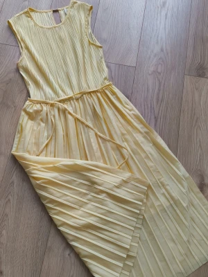 Ljusgul plisserad klänning - Säljer en härlig och sval ljusgul klänning med plisserad design och smalt knytband i midjan. Klänningen är ärmlös och har rund halsringning samt en liten gulddetalj i nacken. Perfekt för sommarens alla tillfällen!