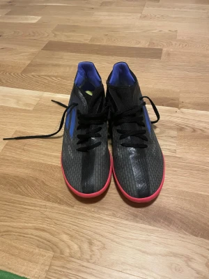 Svarta Adidas futsal skor - Säljer ett par snygga Adidas fotbollsskor i svart med blå detaljer och rosa sula. Skorna har klassisk snörning, låg profil och är tillverkade i syntetmaterial. Perfekta för dig som vill ha stilrena och funktionella skor på planen.