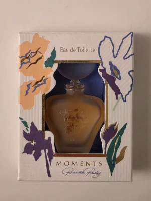 Moments Eau de Toilette Priscilla Presley - Moments Eau de Toilette från Priscilla Presley i en elegant, hjärtformad glasflaska med blommig relief. Förpackningen är dekorerad med färgglada blommor i lila, orange och guld. Innehåller 3 ml doft och kommer i originalkartong.