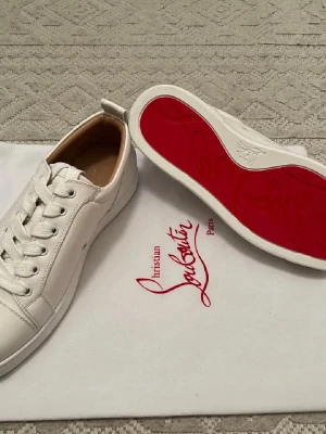 Vita sneakers från Christian Louboutin - Säljer ett par vita sneakers från Christian Louboutin med den ikoniska röda sulan och diskret logga på sidan. Skorna har klassisk låg profil, vita skosnören och är tillverkade i slätt läder med beige innersula. Perfekta för dig som gillar exklusiv och stilren design.