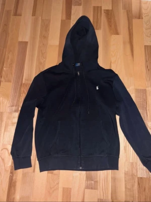 Polo Ralph Lauren hoodie - Säljer en svart hoodie från Polo Ralph Lauren med dragkedja framtill och klassisk vit logga på bröstet. Hoodien har huva med dragsko och två fickor framtill. Tillverkad i mjukt bomullsmaterial som är skönt att ha på sig.