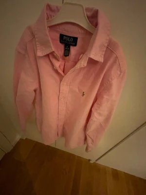 Rosa skjorta från Polo Ralph Lauren - Använd två gånger bara Säljer en klassisk rosa skjorta från Polo Ralph Lauren i storlek M. Skjortan har lång ärm, knäppning framtill och den ikoniska broderade loggan på bröstet. Tillverkad i mjukt bomullstyg och har en snygg krage som ger en clean look.