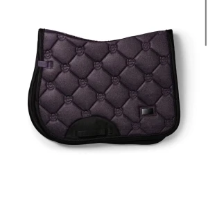 Mörklila schabrak med mönster - Säljer ett mörklila schabrak med quiltat mönster och svarta kanter. Schabraket har små broderade detaljer i rutorna och en praktisk rem för att fästa det på sadeln. Perfekt för dig som vill ha en snygg detalj till hästutrustningen. Pris diskuteras 