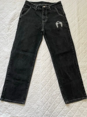 Svarta jeans med broderad ängel - Säljer ett par svarta raka jeans med vit kontrastsöm och en broderad ängel på ena fickan fram. Jeansen har hög midja och klassisk femficksmodell. Materialet är kraftig denim i bomull. Perfekta för dig som gillar streetwear och unika detaljer.