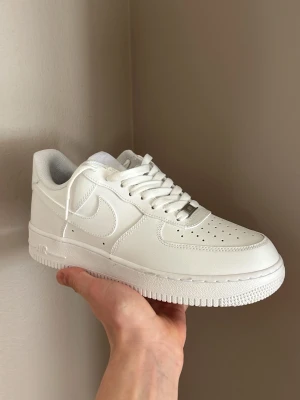 Nike Air Force 1 vita låga sneakers - Klassiska Nike Air Force 1 sneakers i helvitt läder med perforerad tåbox, vita skosnören och diskret Nike-logga på sidan och hälen. Snygg och tidlös modell som passar till det mesta. Låg profil och robust sula för skön komfort.