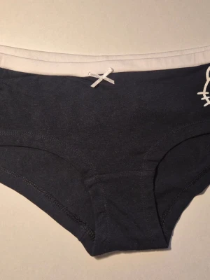 Svarta Hello Kitty trosor med vit rosett stlk M  - Säljer ett par svarta Hello Kitty trosor med vit resårkant och en liten vit rosett framtill. Trosorna har en klassisk hipster-modell och är tillverkade i mjukt bomullsmaterial. Diskret vit detalj på sidan ger en söt touch. Helt oanvända såklart, kommer från ett restparti. Detta par har inga orginalprislappar kvar, men det har många av mina andra annonser. 20% bundlerabatt om du köper flera.