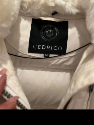 Vit dunjacka med päls från Cedrico - Säljer en lång, vit dunjacka från Cedrico i storlek XS. Jackan har en stor huva med fluffig päls, dragkedja och knappar framtill samt flera fickor med dragkedja. Perfekt för kalla vinterdagar och riktigt snygg design.
