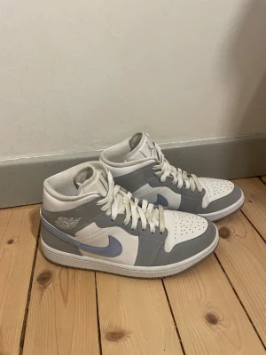 Nike Air Jordan 1 Mid grå/vit/ljusblå - Snygga Nike Air Jordan 1 Mid sneakers i vitt och ljusgrått med lila swoosh. Klassisk siluett med snörning, perforerad tå och rund tå. Perfekta för dig som vill ha en stilren och trendig look.                            Skorna är knappt använda då jag köpte för liten storlek                                                Nypris: 3000 kr 