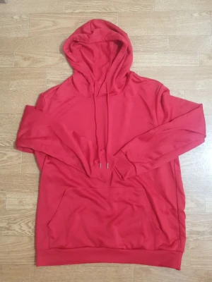 Röd hoodie med magficka - Snygg röd hoodie med stor huva och dragsnören. Klassisk magficka framtill och ribbade muddar vid ärmslut och nederkant. Perfekt för chill dagar eller när du vill ha en enkel och stilren look. Materialet känns mjukt och bekvämt.