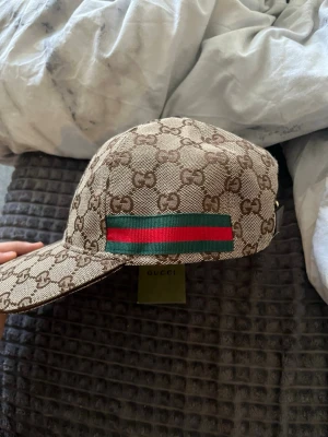 Gucci beige keps med GG-mönster - Säljer en beige keps från Gucci med det klassiska GG-monogrammet över hela kepsen. På sidan finns en grön och röd randig detalj. Justerbar rem bak i brunt läder och insida i ljus textil. Perfekt för dig som vill ha en ikonisk accessoar.