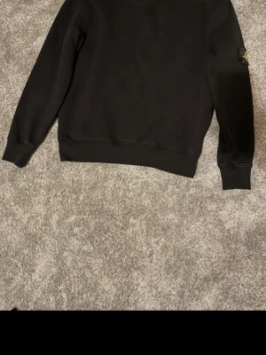 Svart sweatshirt från Stone Island - Säljer en svart sweatshirt från Stone Island med klassisk logga på ärmen. Tröjan har rund halsringning, ribbade muddar och är tillverkad i mjukt bomullsmaterial. Perfekt för en stilren och avslappnad look.
