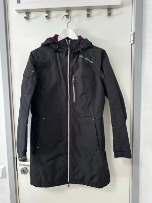 Helly Hansen jacka - Jackan har en dragkedja framtill och en huva. Det finns två fickor med dragkedja i midjehöjd och en mindre ficka med dragkedja på bröstet. Ärmsluten är justerbara med kardborreband.