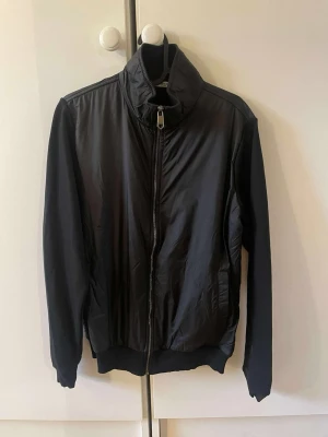 Svart bomberjacka från Givenchy - Svart bomberjacka från Givenchy med hög krage och dragkedja framtill. Jackan har broderad Givenchy Paris-logga i rött på ryggen och ribbade muddar vid ärmslut och midja. Tillverkad i glansigt syntetmaterial för en modern look.