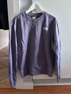 Lila sweatshirt från The North Face - Säljer en lila sweatshirt från The North Face med klassisk logga på bröstet. Tröjan har rund halsringning, ribbade muddar och är tillverkad i mjukt bomullsmaterial. Perfekt för en avslappnad och stilren look.