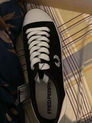 Svarta Fred Perry sneakers låga - Svarta sneakers från Fred Perry med vit sula och vita skosnören. Klassisk låg modell med broderad logga på sidan och ventilerande metallhål. Ovandelen är i canvas och sulan i gummi. Perfekta för en clean och stilren look.