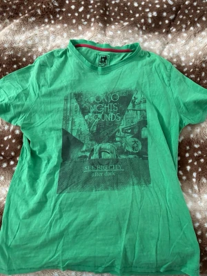 Grön t-shirt Tokyo Nights från Red Sew - Grön t-shirt med tryck 'Tokyo Nights Sounds' och stadsmotiv framtill. Rund hals med röd detalj på insidan. Perfekt för dig som gillar streetstyle och grafiska prints.