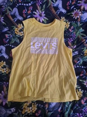 Gul ärmlös linnetopp från Levi's - Snygg gul ärmlös linnetopp från Levi's med ett vitt grafiskt tryck på bröstet. Toppen har rund halsringning och är gjord i mjuk bomull, perfekt för en avslappnad och cool stil.