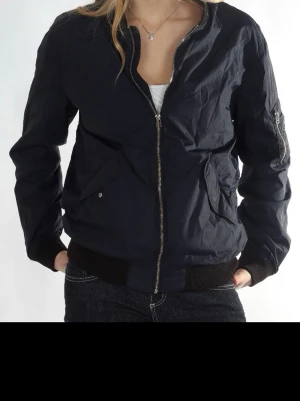 Svart bomberjacka med dragkedja - Snygg svart bomberjacka med silverfärgad dragkedja framtill och ribbade muddar vid ärmslut och midja. Jackan har fickor med knappstängning och en klassisk ficka på ärmen. Perfekt för en cool och avslappnad stil.