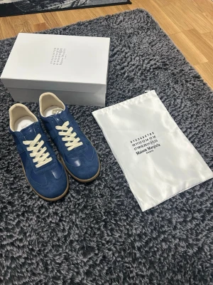 Blå Maison Margiela Replica sneakers - Maison Margiela Replica sneakers i blå mocka och läder med vita snören och beige innersula. Klassisk låg modell med gummisula och diskreta detaljer. Kommer med originalkartong och dustbag. Perfekta för dig som gillar stilrena och tidlösa sneakers.