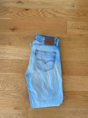 Ljusblå Levi's jeans  - Säljer ett par klassiska ljusblå Levi's jeans med rak passform och fem fickor. Jeansen har den ikoniska röda Levi's-lappen på bakfickan och en brun läderpatch i midjan. Tillverkade i bomull med snygga slitningar framtill för en avslappnad look.