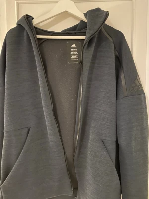 Mörkgrå hoodie från Adidas - Snygg mörkgrå hoodie från Adidas med dragkedja framtill och två stora fickor. Adidas-logga på ärmen och huva för extra stilpoäng. Tillverkad i ett mjukt och bekvämt material som passar perfekt till en avslappnad look.