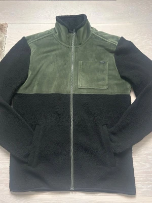 Grön och svart fleecejacka från Bula - Säljer en riktigt snygg och unik pilejacka från det norska märket Bula. Jackan har en cool design med olivgrön manchester (corduroy) över axlar och bröst, samt svart, mjuk teddyfleece på resten. Perfekt för både stadshäng och friluftsliv! • Märke: Bula • Storlek: S (Small) • Skick: Mycket bra skick, sparsamt använd. • Detaljer: Bröstficka med logga, hellång dragkedja och hög krage som värmer gott. • Passform: Normal i storleken, skön stretch och mjuk insida. En perfekt jacka för våren #fleece