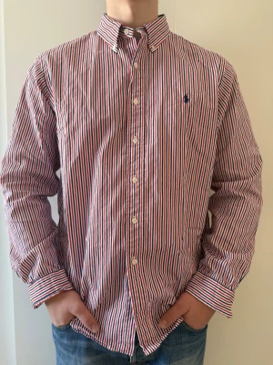Randig skjorta från Ralph Lauren - Klassisk skjorta från Ralph Lauren med röd, blå och vit randigt mönster. Riktigt bra skick och funkar perfekt för layering eller bara ha som den är, storlek XL custom fit