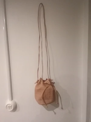 Beige bucket bag i mocka - Snygg beige axelväska i bucket-modell med lång axelrem och dragskostängning. Väskan är gjord i mjuk mocka och har en minimalistisk design utan synliga loggor eller detaljer. Perfekt för dig som gillar enkel och stilren look.
