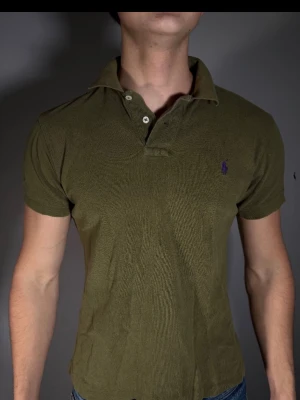 Olivgrön pikétröja från Ralph Lauren - Klassisk olivgrön pikétröja från Ralph Lauren med korta ärmar och två knappar vid kragen. Tröjan har den ikoniska lilla lila logotypen broderad på bröstet. Perfekt till jeans eller shorts för en avslappnad men snygg look.